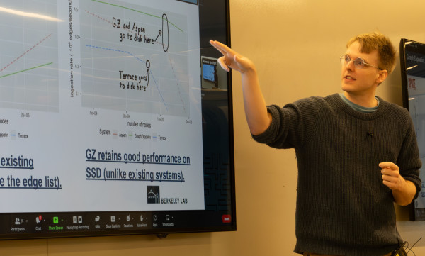 A man with a fair complexion and light brown hair wearing glasses and a casual sweater points to a scientific graph displayed on a large monitor.